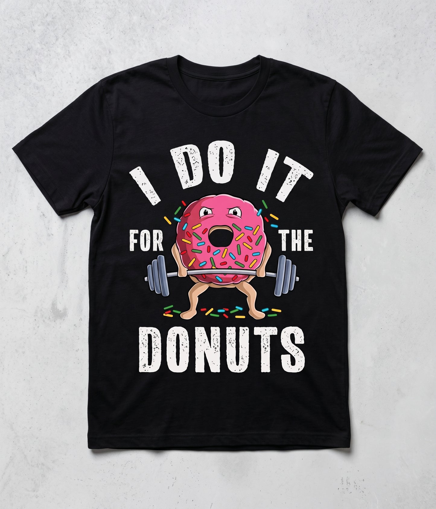 A black short-sleeve t-shirt with a graphic print of a cartoon pink donut lifting a barbell, surrounded by the text 'I DO IT FOR THE DONUTS' in white distressed lettering, displayed on a light gray textured background.