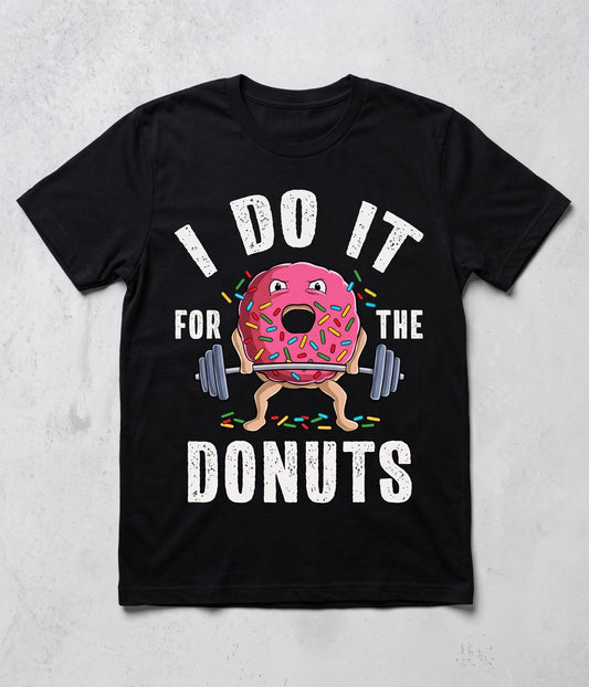 A black short-sleeve t-shirt with a graphic print of a cartoon pink donut lifting a barbell, surrounded by the text 'I DO IT FOR THE DONUTS' in white distressed lettering, displayed on a light gray textured background.
